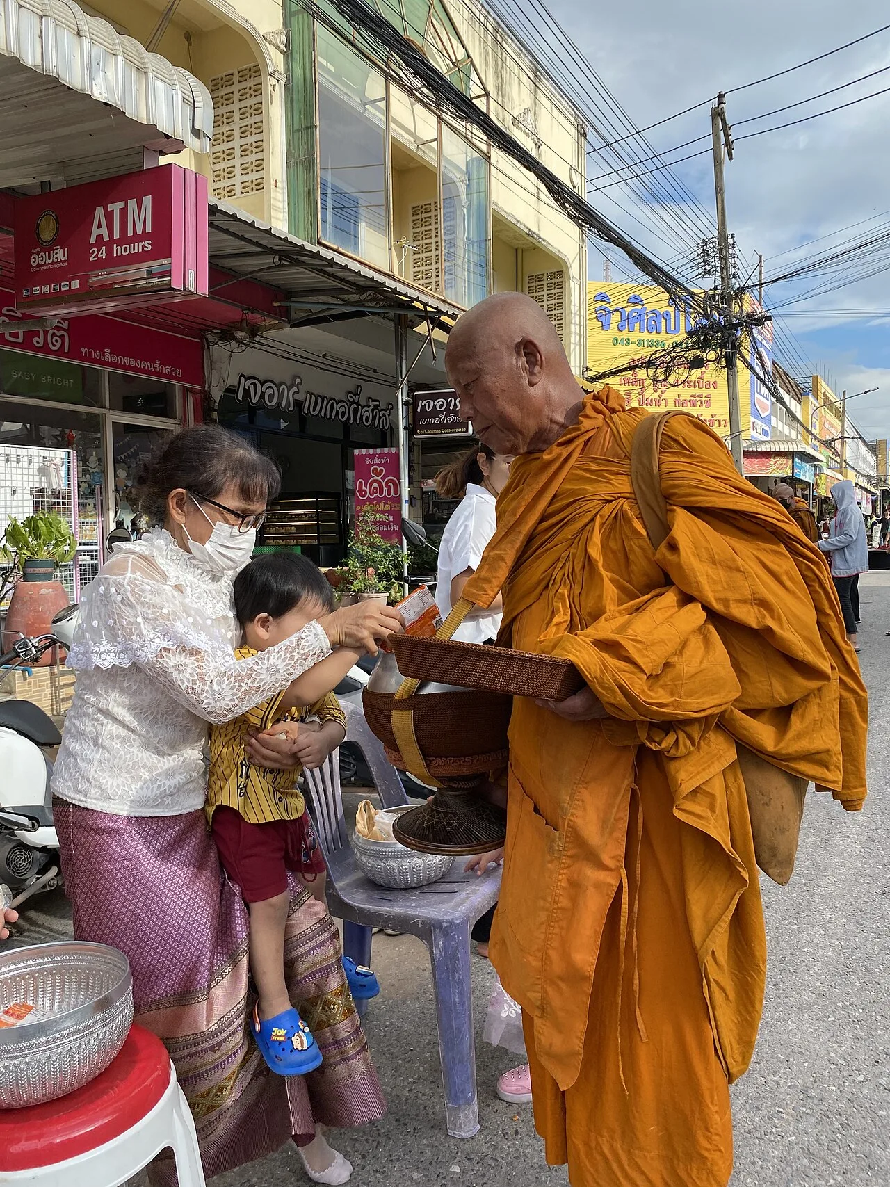 ผู้ใหญ่ช่วยประคองเด็กขณะใส่ของลงในบาตรพระบริเวณหน้าร้านในชุมแพ