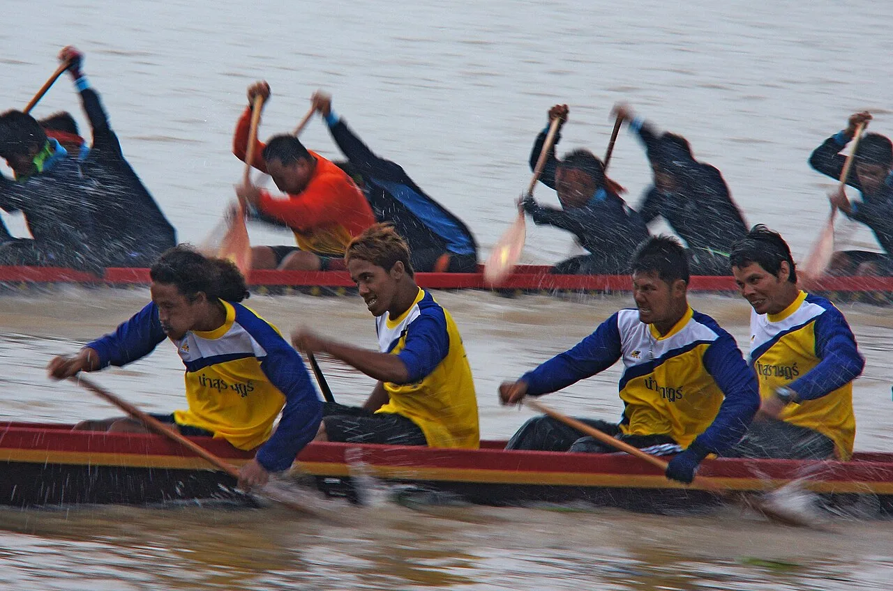 นักกีฬาสองทีมพายเรือยาวขนานกันในแม่น้ำ มีละอองน้ำกระเซ็นจากฝีพาย