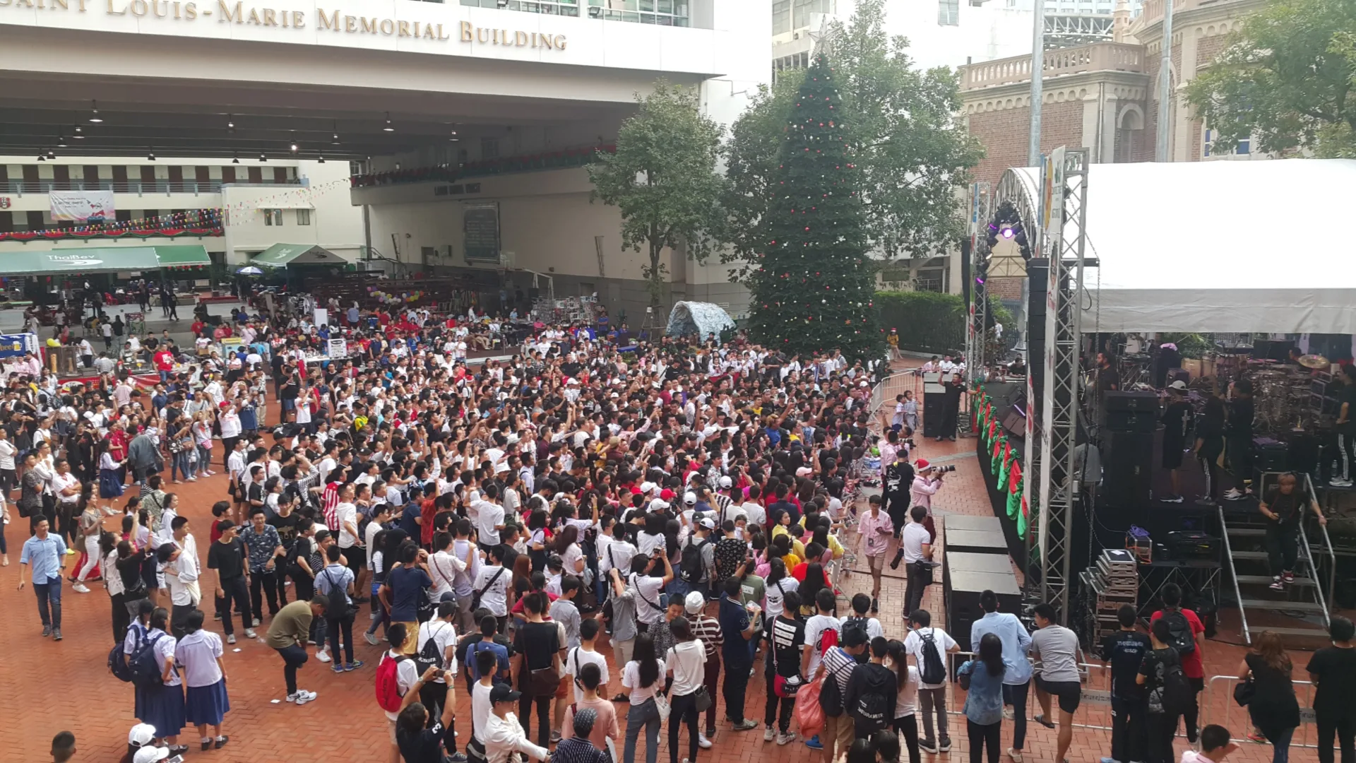 ผู้ชมจำนวนมากยืนหน้าด้านข้างเวทีคอนเสิร์ตกลางแจ้งในลานอาคาร