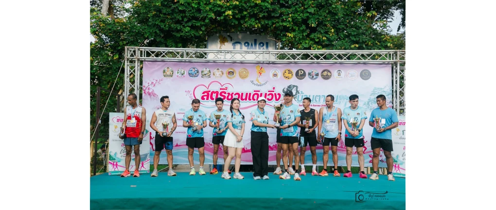 ผู้ร่วมกิจกรรมถือถ้วยบนเวทีหน้าป้ายสตรีชวนเดิน-วิ่ง บ้านตาขุน จากภาพลำดับสองในรายงานเทศบาลตำบลบ้านตาขุน
