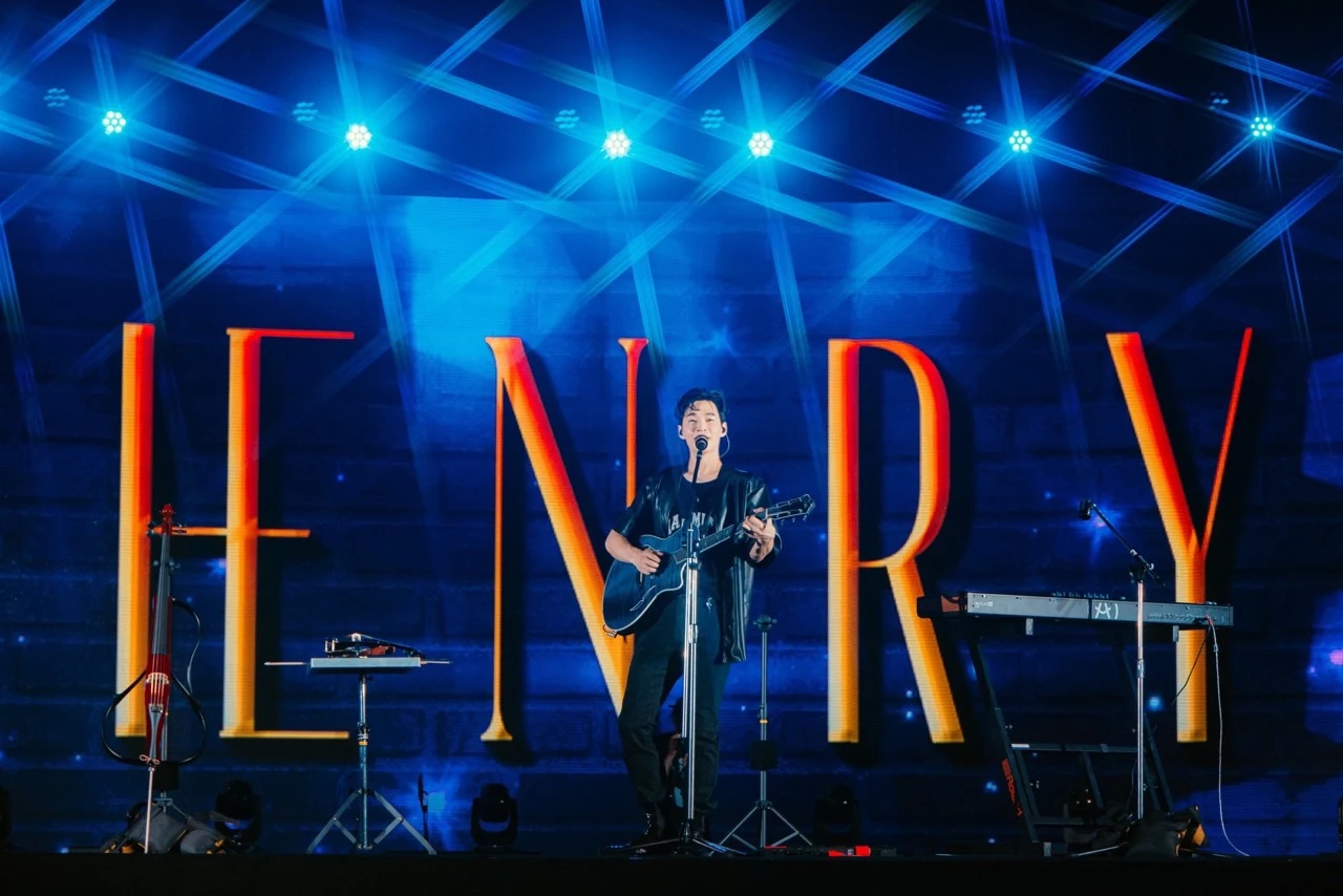 นักดนตรีถือกีตาร์หน้าไมโครโฟนบนเวทีแสงสีฟ้า มีตัวอักษร HENRY ขนาดใหญ่ด้านหลัง