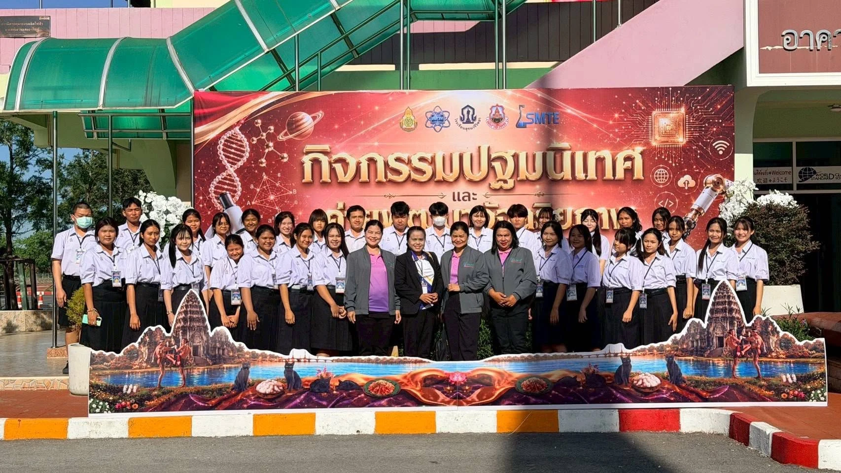ผู้ร่วมกิจกรรมยืนถ่ายภาพหมู่หน้าป้ายค่าย SMTE และอาคารที่มีหลังคาทางเดินสีเขียว