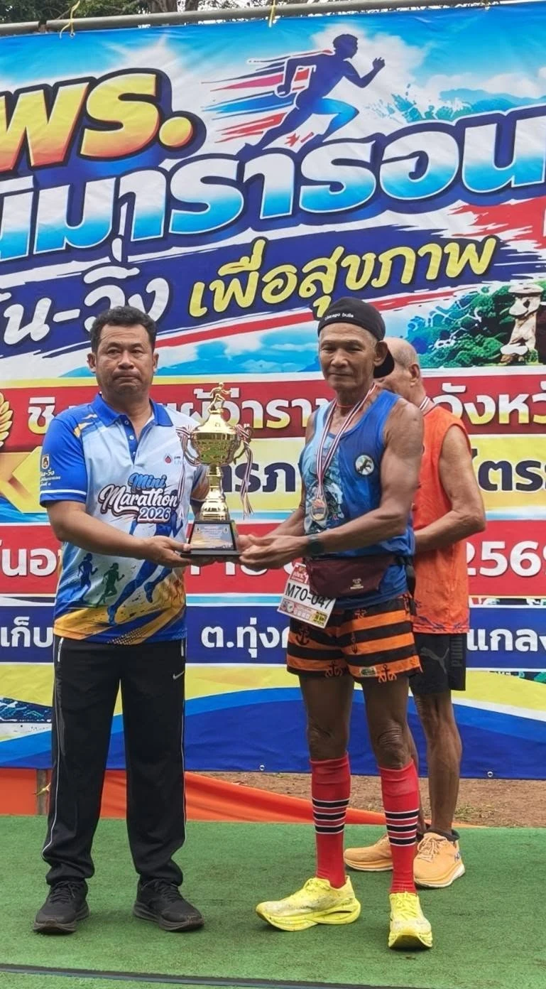 ผู้ร่วมงานถือถ้วยรางวัลบริเวณป้าย อปพร. มินิมาราธอน จากชุดภาพของเพจงาน