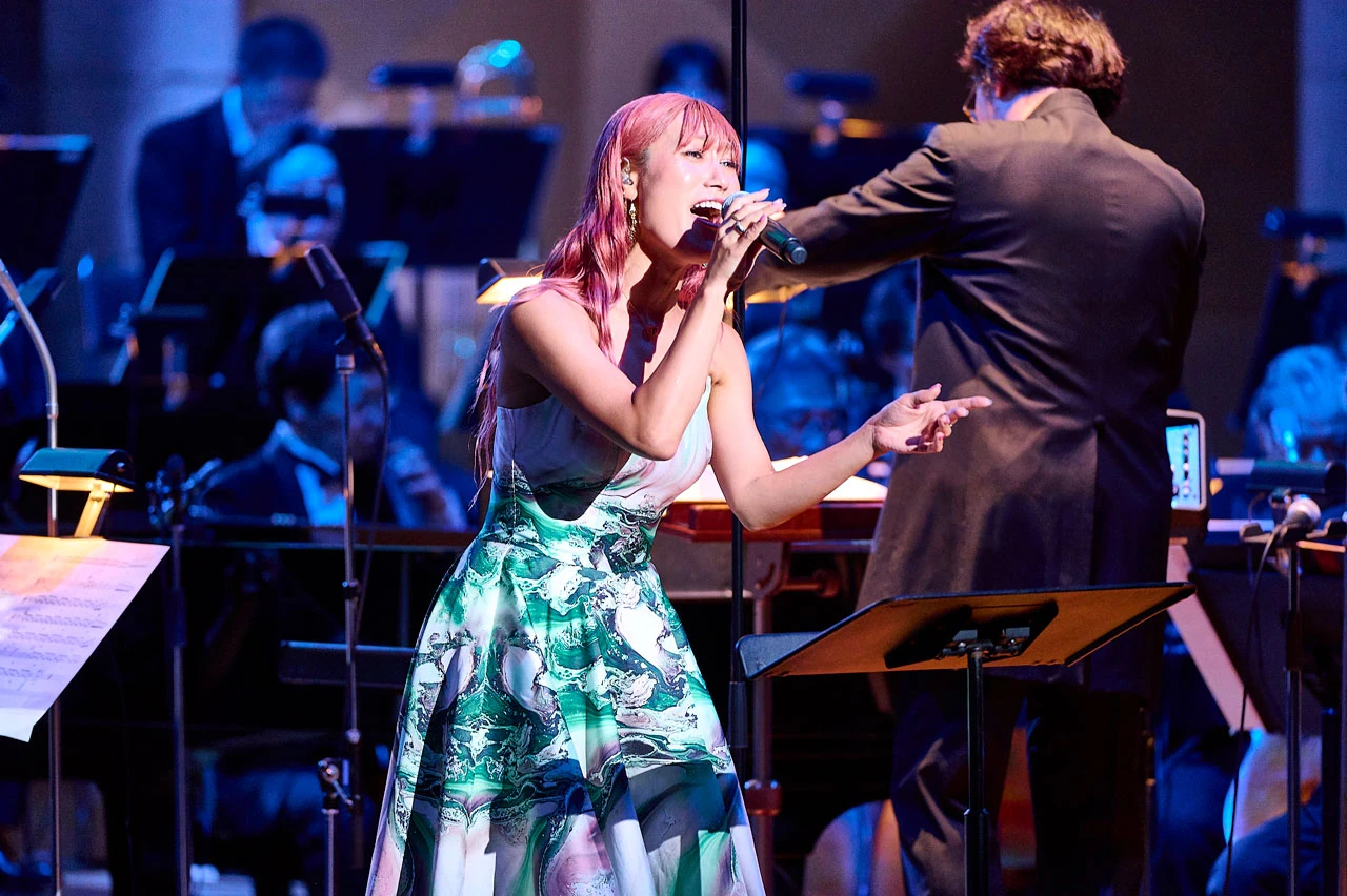 ภาพนักร้องถือไมโครโฟนร่วมกับวงออร์เคสตราในโปรแกรม Metaphor: ReFantazio Orchestra Concert จากแกลเลอรีทางการ ไม่ระบุวันที่ถ่ายและสถานที่