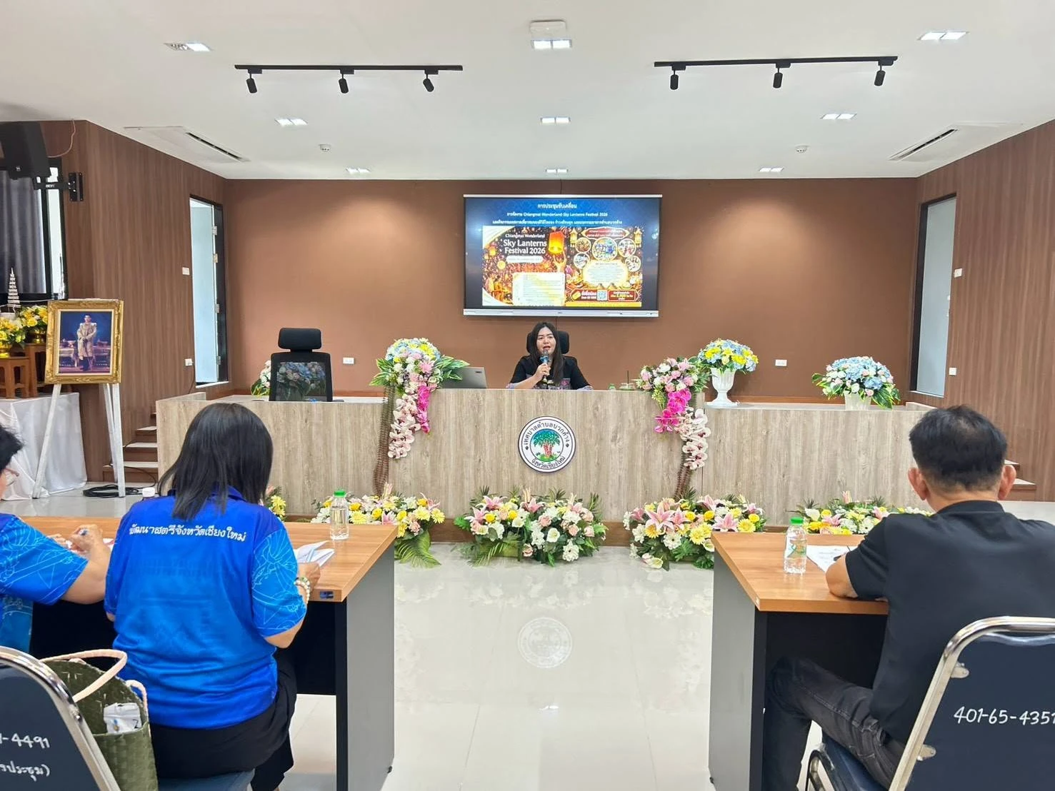 ผู้พูดถือไมโครโฟนและผู้ฟังในห้องประชุมเทศบาลตำบลบวกค้าง พร้อมจอชื่อ Chiangmai Wonderland 2026 ภาพเตรียมงาน