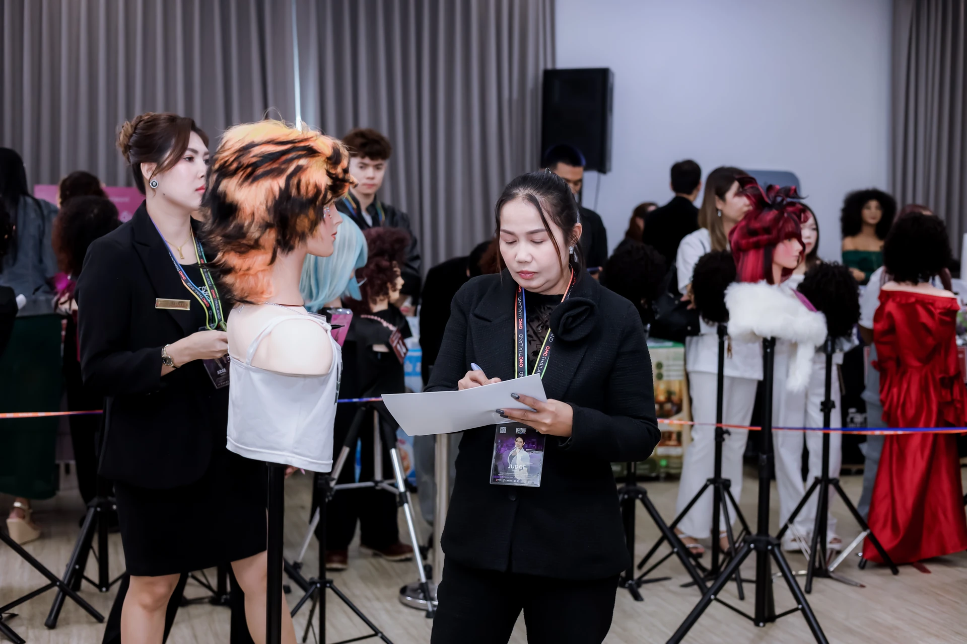 กรรมการถือเอกสารตรวจผลงานใกล้หุ่นทรงผมสีส้มและหุ่นอื่น ๆ ใน BEAUTY CULTURE FESTIVAL กรุงเทพฯ ปี 2569 ภาพเทศกาลโดยรวม