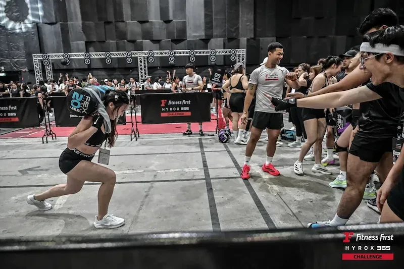 ผู้เข้าแข่งขันก้าวย่อขาพร้อมแบกถุงน้ำหนัก โดยมีผู้ชมเชียร์ในงาน Fitness First X HYROX365 Challenge ปี 2568
