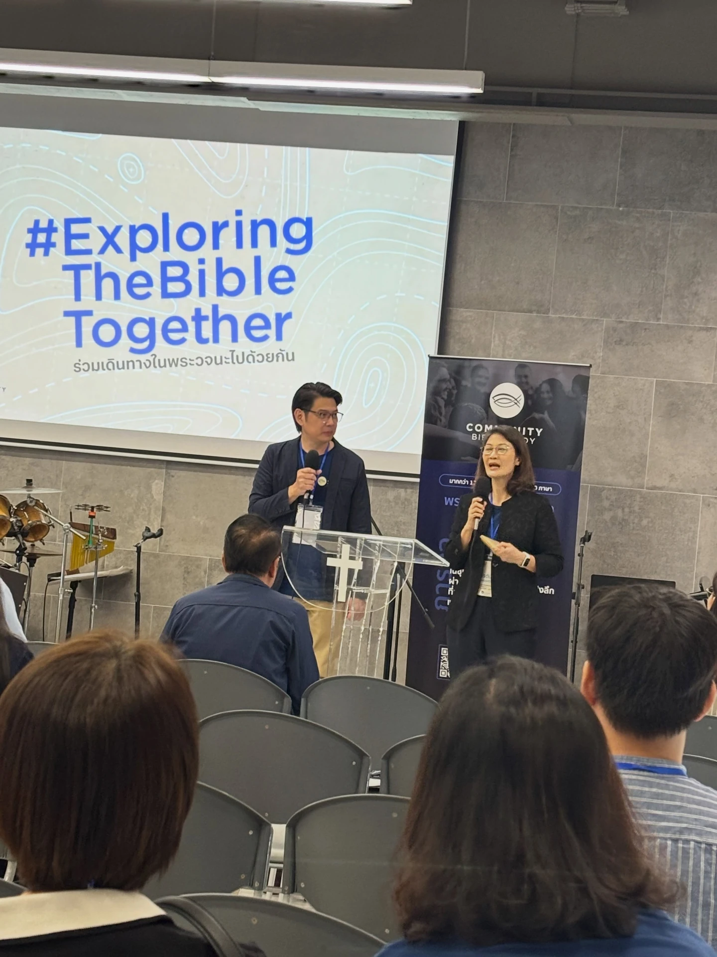 ผู้พูดถือไมโครโฟนช่วงเปิดงาน Day 1 และผู้ร่วมงาน CBS Thailand ปี 2569 พร้อมจอ Exploring the Bible Together