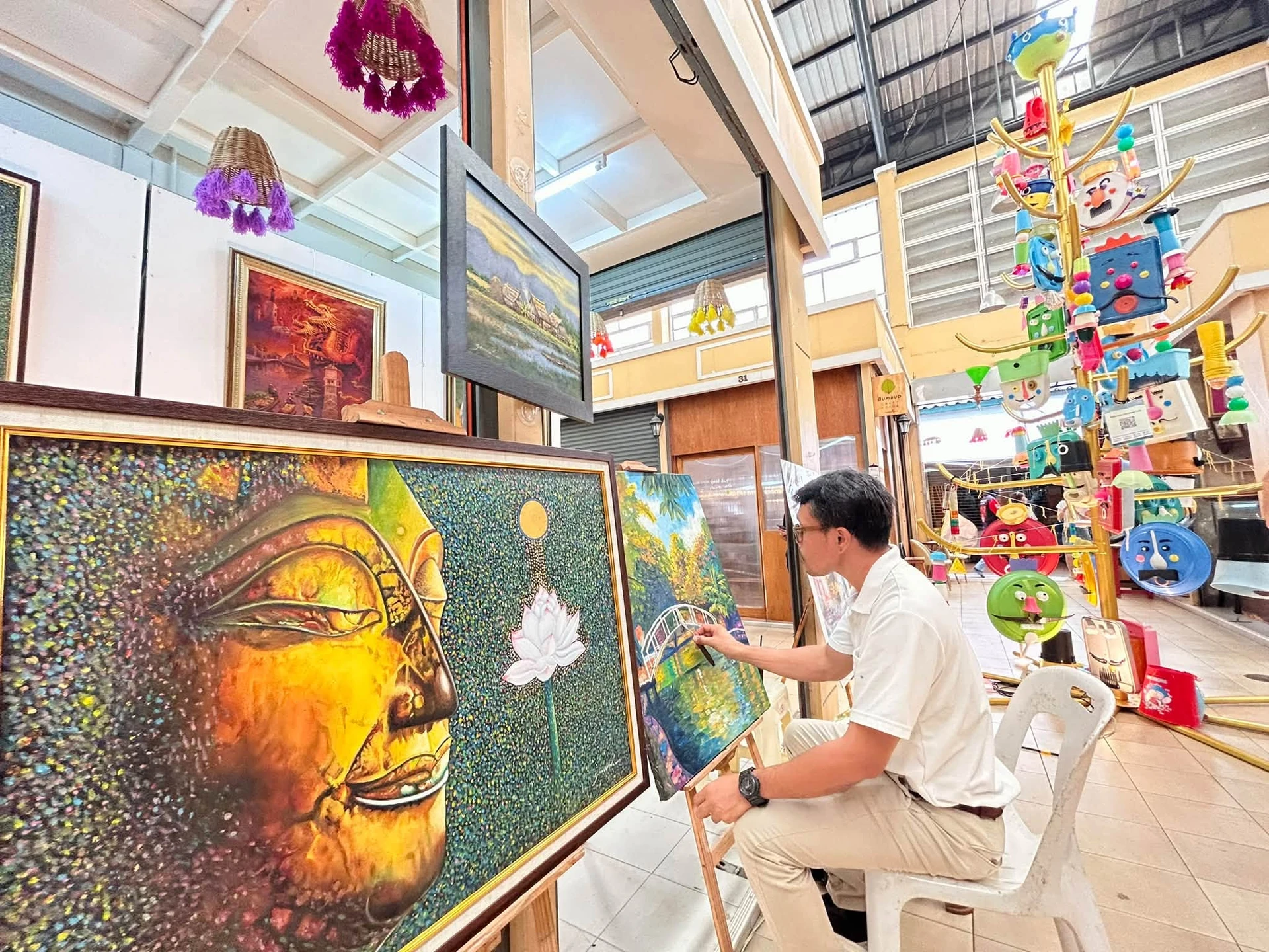 ผู้วาดภาพนั่งทำงานข้างภาพวาดและชิ้นงานศิลปะ ภายในงานเมืองเก่า เล่าเรื่องอาร์ต ราชบุรี ปี 2569 ไม่ระบุบุคคลจากใบหน้า